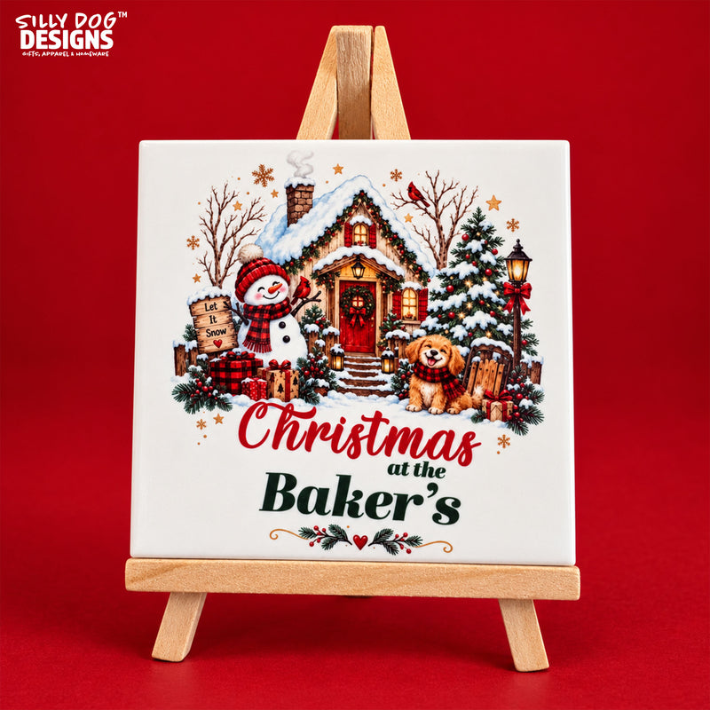 A square ceramic tile with a festive Christmas illustration, displayed on a small wooden easel against a solid red background. The tile features a snow-covered cottage, a snowman, a dog, and the text 'Christmas at the Baker's'.