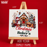 A square white ceramic Christmas canvas print displayed on a small wooden easel against a solid red background. The print features a festive illustration of a snow-covered cottage, a snowman, a dog, a Christmas tree, and the customizable text 'Christmas at the Baker's' with a floral accent.
