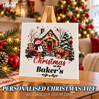 A personalized Christmas tile displayed on a wooden stand, featuring a festive illustration of a snowman, dog, and bakery scene with the text 'Christmas at the Baker's'. The background shows a cozy, blurred Christmas living room with a decorated tree and warm lights.