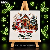 A square ceramic tile displayed on a small wooden easel, featuring a festive Christmas village scene with a snowman, dog, house, tree, and the text 'Christmas at the Baker's'. The background is black with glowing green holiday decorations, and the Silly Dog Designs logo appears in the top left.