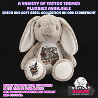 A plush bunny toy centered on a black background with promotional text. The bunny is light gray with cream inner ears and a white belly patch printed with a tattoo-style design reading 'Junkie and Rehab.