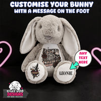 A plush gray bunny toy with long floppy ears sits against a black background with pink heart-shaped neon glow accents. Large white text at the top reads 'CUSTOMISE YOUR BUNNY WITH A MESSAGE ON THE FOOT,' and the bunny has printed sample designs on its belly and paws, including a tattoo-style skull-and-sword graphic and the name 'LEONIE.