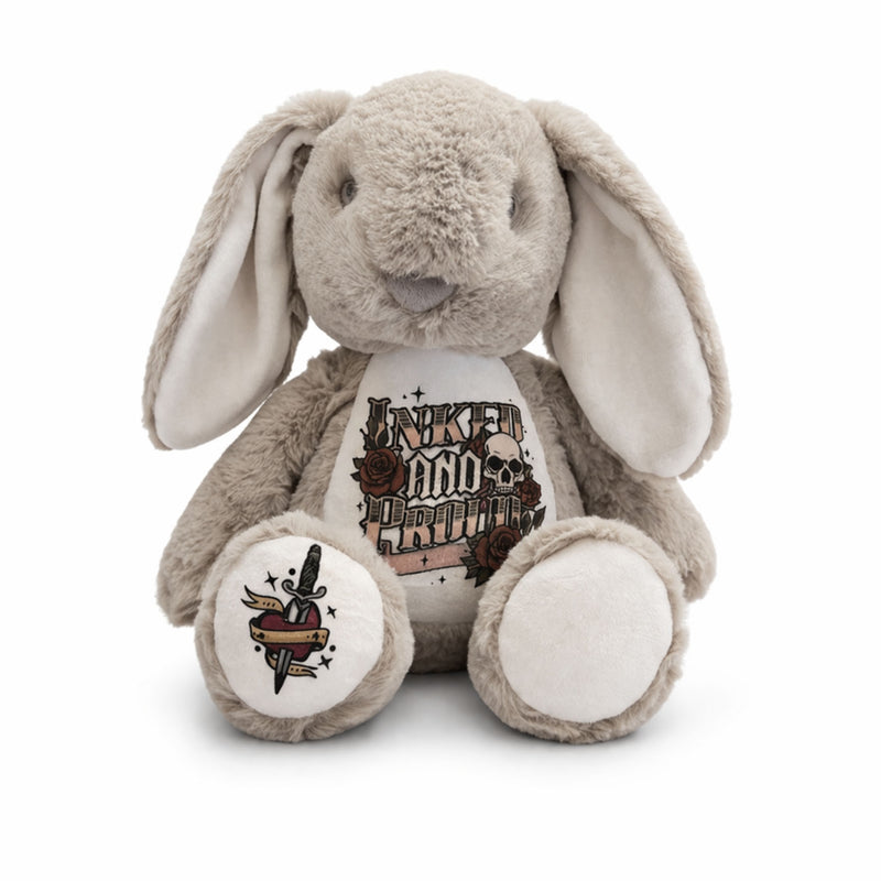A plush bunny toy in light gray and cream, featuring a white belly patch with a tattoo-style graphic that reads 'INKED AND UNLEASHED' with roses and a dagger, and a matching graphic on one paw.