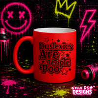 A bright red ceramic mug with black text reading 'Dyslexics Are People Poo' surrounded by small stars, shown on a dark background with neon spray-paint splatters and paint cans. A 'SILTY DOG DESIGNS' logo appears in the bottom right corner.