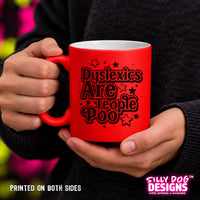 A person holding a bright red ceramic mug with black text that reads 'Dyslexics Are People Poo' surrounded by small stars. The person is wearing a dark gray sweater, and the background is a blurred multicolor outdoor setting.