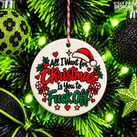 A round white ceramic Christmas ornament with a humorous message, hanging on a green Christmas tree with lights and ornaments. The ornament features a Santa hat, holly, candy canes, and the text 'All I Want for Christmas Is You to Fuck Off' in red and green lettering.