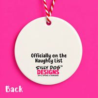 A round white ceramic ornament or tag hanging from a red-and-white striped string on a bright pink background. The front has printed text reading 'Officially on the Naughty List' and 'SILLY DOG DESIGNS GIFTS, APPAREL & HOMEWARE.