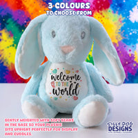 A light blue plush bunny toy with long floppy ears, a white belly patch that reads 'Welcome to the world' with small heart and star graphics, and a colorful rainbow paint-splatter background. Text at the top says '3 COLOURS TO CHOOSE FROM,' and the Silly Dog Designs logo appears at the bottom right.
