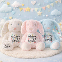 Three plush bunny toys in white, pink, and blue with 'welcome to the world' text on a soft surface.