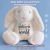 A white plush bunny toy with long floppy ears sits against a light blue background. The front has a printed message reading 'welcome to the world' with small colorful hearts and stars.