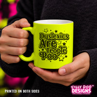 A person holding a bright neon yellow-green ceramic mug with black text that reads 'Dyslexics Are People Poo' surrounded by small stars. The person is wearing a dark charcoal sweater, and the background is a soft, blurred pink and green outdoor setting.
